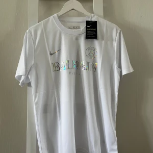 Vit Nike x Balmain Paris t-shirt - Snygg vit t-shirt från Nike i samarbete med Balmain Paris. T-shirten har holografiskt tryck framtill med Balmain-logga och Paris-märke. Klassisk rund hals, korta ärmar och tillverkad i lätt polyester som andas. Perfekt för dig som gillar exklusiva samarbeten och sportig stil.