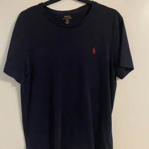 Mörkblå t-shirt från Polo Ralph Lauren - Snygg mörkblå t-shirt från Polo Ralph Lauren med klassisk röd broderad logga på bröstet. Modellen är kortärmad och i 100% bomull, vilket gör den riktigt skön att bära. Perfekt basic-plagg med stilren design och rund halsringning.