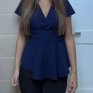 Mörkblå v-ringad blus - Snygg mörkblå blus med v-ringning och korta ärmar. Toppen har markerad midja och ett utsvängt, peplumliknande avslut som ger en feminin siluett. Perfekt för dig som gillar stilrena och klassiska plagg med en modern twist.