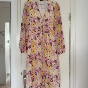 Blommig långklänning från Gina Tricot - Superfin långklänning med blommigt mönster i rosa, lila och gult. Klänningen har v-ringning, knappar framtill och långa ärmar i ett lätt, luftigt material. 