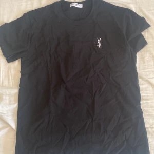 Svart Saint Laurent t-shirt med logga - Svart t-shirt från Saint Laurent med klassisk passform och korta ärmar. På bröstet finns en broderad YSL-logga i vitt. Tillverkad i mjuk bomull och har en rund halsringning. Perfekt för en clean och stilren look.