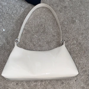 Vit axelväska i glansigt material - Snygg vit axelväska med stilren och minimalistisk design. Väskan har en glansig finish och en enkel axelrem. Perfekt storlek för det viktigaste och stängs med dragkedja upptill. Passar dig som gillar en clean och modern look. Den är från H&M 