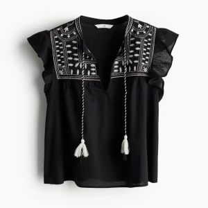 Svart blus med broderi och volang - Svart blus från H&M med vita broderier upptill och snygga volangärmar. Blusen har knytband med tofsar framtill och en avslappnad passform. Perfekt för dig som gillar boho-stil och detaljer.