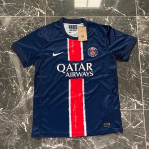 PSG Dembele 10 matchtröja Nike - Paris Saint-Germain fotbollströja med O. Dembélé och nummer 10 på ryggen. Djupblå bas med röd och vit vertikal rand på framsidan, PSG-emblem på bröstet och Qatar Airways-tryck. Tillverkad i lätt och ventilerande material, perfekt för match eller träning.