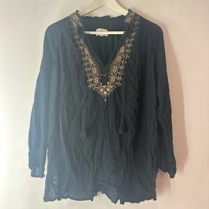 Svart broderad blus från Ralph Lauren - Svart blus från Ralph Lauren med v-ringning och snyggt broderi i guld och vitt längs halsringningen. Långärmad och lös passform med tunna snören framtill. Perfekt för dig som gillar boho-vibe och detaljer. Lite släppta trådar i broderiet men inget som är sönder 