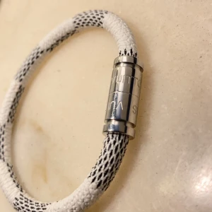 Vitt armband i textil - Snyggt armband i vit textil med svarta detaljer och ett stilrent magnetlås i silverfärgad metall. Armbandet har ett sportigt och modernt utseende med diskret logga ingraverad på låset. Perfekt accessoar för att lyfta din outfit.