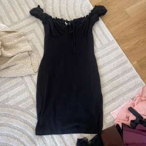 Svart offshoulder kortklänning - Snygg svart kortklänning med offshoulder och volangdetaljer upptill. Klänningen har en smickrande passform och knytband vid bysten. Perfekt för dig som gillar en enkel men trendig stil.