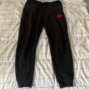 Svarta mjukisbyxor från Hugo - Svarta mjukisbyxor från Hugo med röd logopatch på vänster ben. Byxorna har dragsko i midjan, ribbade muddar vid benslut och är gjorda i mjuk bomullsmix. Perfekta för chill dagar eller när du vill vara bekväm men ändå stilren.