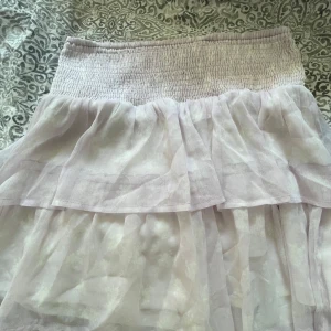 Ljuslila volangkjol från Vero Moda - Supersöt ljuslila kortkjol från Vero Moda med bred smock i midjan och dubbla volanger. Kjolen är luftig och har en lätt transparent känsla, perfekt för varma dagar. Styla med en enkel topp och sneakers för en chill look.