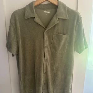 Olivgrön frottéskjorta från Kappahl - Olivgrön skjorta i mjuk frotté från Kappahl. Skjortan har korta ärmar, klassisk krage, knäppning framtill och en bröstficka. Perfekt för en chill och avslappnad stil under sommaren. Aldrig använd - nyskick. Nypris 399