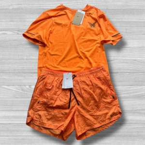 Nike Running Division - Nike Running Divison shortsset i orange färg. Tillverkad i lätt och snabbtorkande syntetmaterial, perfekt för träning. Diskret Nike-logga på bröstet och Dri-FIT-tryck på ryggen. Sportig och clean look. T-shirt storlek: S. Shorts storlek: L