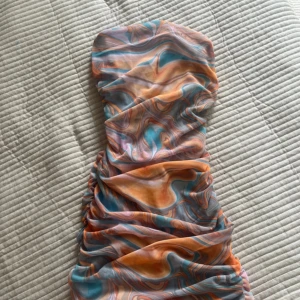 Färgglad axelbandslös kortklänning - Säljer en superfin axelbandslös kortklänning med swirlmönster i orange, blått och rosa. Klänningen har en tight passform och snygga draperingar framtill. 
