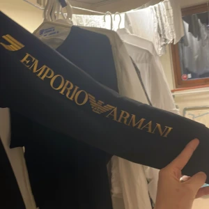Svart Emporio Armani EA7 tjocktröja  - Svart vindjacka från Emporio Armani EA7 med guldfärgad logga på bröstet och stort tryck längs ärmen. Tröjan har huva och hel dragkedja framtill. Snygg och sportig look som sticker ut med exklusiva detaljer.