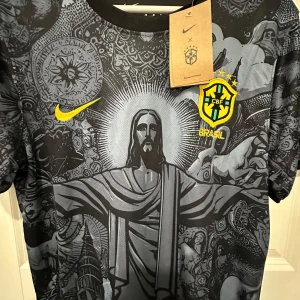 Nike Brasil CBF svart grafisk fotbollströja - Unik svart Brasilien CBF fotbollströja från Nike med detaljerat grafiskt tryck av Kristusstatyn och brasilianska symboler. Gula loggor och broderat klubbmärke på bröstet. Tillverkad i lätt, ventilerande funktionsmaterial. Rund hals och korta ärmar.