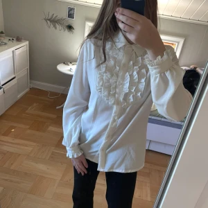 Vit blus med volangdetaljer - Säljer en vit blus med långa ärmar och snygga volangdetaljer framtill. Skjortan har klassisk krage, knäppning hela vägen och volanger även vid ärmsluten. Perfekt för dig som gillar en romantisk och trendig look.💞står ingen storlek men skulle gissa på S😊