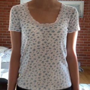 Blommig topp med spets från Monki - Supersöt topp från Monki i vitt med ljusblått blommönster. Den har korta ärmar, ribbad struktur och en fin spetskant runt den djupa, rundade urringningen. 