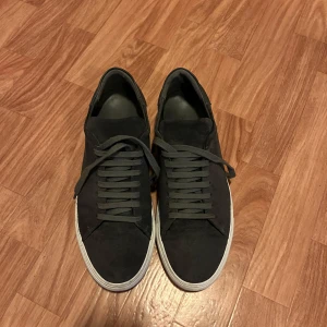 Arigato skor  - Stilrena grå sneakers från Axel Arigato i mjuk mocka med vita platta sulor. Skorna har rund tå, diskret logga på sidan och klassisk snörning. Perfekta för dig som gillar clean stil.