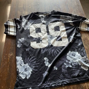 Svart blommig t-shirt Black Squad - Cool svart t-shirt från Black Squad med stora vita siffror '99' på bröstet och blommigt mönster i grått och vitt. Ärmarna har sportiga ränder och t-shirten har en avslappnad passform. Perfekt för dig som gillar streetstyle och unika prints.