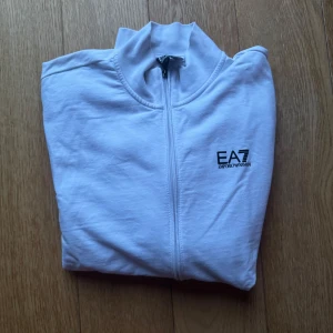 Vit zipjacka från EA7 Emporio Armani - Snygg vit jacka från EA7 Emporio Armani med hel dragkedja och diskret logga på bröstet. Jackan har en clean look, ståkrage och är tillverkad i mjukt bomullsmaterial. Perfekt för dig som gillar stilren och sportig vibe.