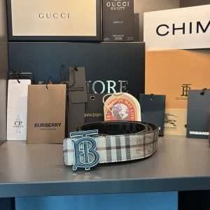 Burberry bälte (äkta) - Snyggt Burberry-bälte med klassiskt rutigt mönster i beige, brunt och rött. Bältet har ett stort metallspänne med Burberrys ikoniska B-logga och är tillverkat i skinn. Perfekt accessoar för att lyfta din outfit med en lyxig touch.