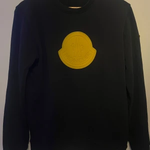 Moncler - Snygg svart sweatshirt från Moncler med stor orange logga på bröstet - riktigt unik design som sticker ut. Skön passform med ribbade muddar och krage. Gjord i mjuk och lite tyngre bomull, perfekt för både vardags och mer stilrena outfits Även en orange logga på ärmen. Vet tyvärr inte om den är äkta där av priset, men kvaliteten känns bra och den ser riktigt clean ut.