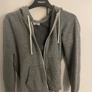 Grå hoodie med dragkedja från Divided - Mysig grå hoodie från Divided med huva, vita snören och dragkedja framtill. Tröjan har känguruficka och ribbade muddar vid ärmslut och nederkant. Perfekt för chill dagar eller när du vill ha en enkel och stilren look.