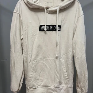 Vit hoodie från American Eagle - Säljer en stilren vit hoodie från American Eagle med broderad logga på bröstet. Tröjan har huva med snörning, stor magficka och långa ärmar. Perfekt för en chill och avslappnad look.