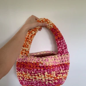 Handgjord  virkad axelväska i rosa orange och gul  - Superfin handgjord axelväska i chunky virkat material med breda ränder i rosa, orange och gult. Väskan har en rundad form och ett rejält handtag som gör den enkel att bära. Perfekt för dig som vill sticka ut med färg och unik stil. Väskan rymmer även de mesta du kan behöva. 