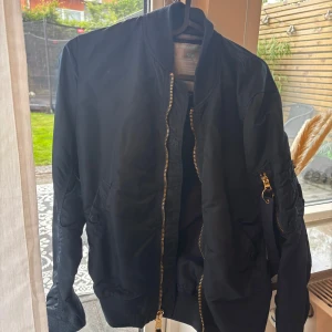 Svart bomberjacka Alpha Industries - Säljer en svart bomberjacka från Alpha Industries i storlek medium. Jackan har klassisk passform, guldiga dragkedjor och fickor på ärmen. Tillverkad i slitstark polyester och perfekt för dig som vill ha en stilren och tidlös look.