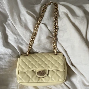 Quiltad axelväska från Guess i ljusgul - Säljer en quiltad beige axelväska från Guess med guldfärgad kedjerem och snyggt spänne framtill. Väskan har flera fack inuti, dragkedja och klassisk design som passar till många outfits. Perfekt storlek för det viktigaste och ger en lyxig känsla.
