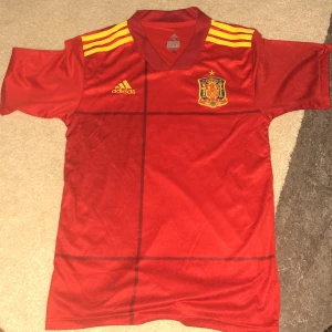 Spaniens röd fotbollströja Adidas - Röd fotbollströja från Adidas med Spaniens landslagsmärke på bröstet och gula detaljer. Tröjan har korta ärmar, gula ränder på axlarna och ett subtilt rutmönster framtill. Perfekt för dig som vill visa ditt stöd för Spanien.