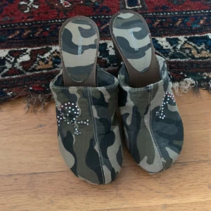 Y2k - Camo Clogs med pirat dödskalle  - Unika träskor med camouflage-mönster i grönt, beige och svart. Ovandelen är dekorerad med en pirat dödskalle. Använd gärna köp nu ❤️