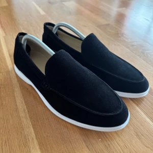 Svarta mockaloafers med vit sula - Stilrena svarta loafers i mocka med vit platt sula. Skorna har en klassisk, minimalistisk design med rund tå och är perfekta för en clean och modern look. Lätta att ta på och passar till både jeans och chinos.