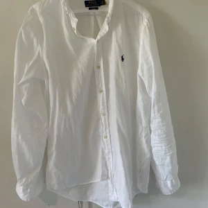 Vit linneskjorta från Polo Ralph Lauren - Klassisk vit skjorta i linne från Polo Ralph Lauren med broderad marinblå logga på bröstet. Skjortan har lång ärm, knappar framtill och en avslappnad passform. Perfekt för en clean och stilren look.