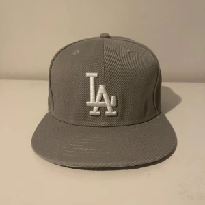 Grå LA Dodgers keps från New Era - Snygg grå keps från New Era med LA Dodgers-logga broderad framtill i vitt. Kepsen har platt skärm, broderad World Series 1988-detalj på sidan och MLB-logga bak. Tillverkad i bomullstyg, perfekt för dig som gillar sportig streetstyle.