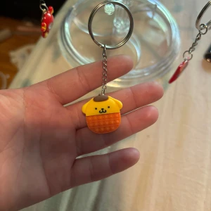 Nyckelring med PomPomPurin-figur - Söt nyckelring med PomPomPurin-figur i gult och orange. Figuren har ett gulligt ansikte, brunt lock och bubblig textur på magen. Perfekt att hänga på väskan eller nycklarna för en extra kawaii touch.