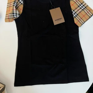 Burberry svart t-shirt med rutiga ärmar - Svart t-shirt från Burberry med klassiska rutiga korta ärmar i beige, rött och svart. T-shirten har rund halsringning och är tillverkad i mjuk bomull. Perfekt för dig som vill ha en stilren look med en touch av lyx.