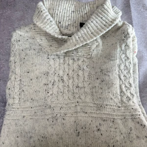 Grå stickad tröja från Chaps - Säljer en mysig grå stickad tröja från Chaps med kabelstickat mönster på framsidan. Tröjan har en hög krage och långa ärmar, perfekt för kyliga dagar. Den är i en neutral grå färg med små mörka prickar som ger en fin textur.