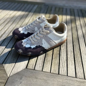 Masion margiela - Snygga sneakers som heter Masion margiela,med ett unikt färgstänk-mönster i svart och beige. Skorna har en klassisk snörning och en bekväm sula. Perfekta för dig som vill sticka ut med en stilren look.