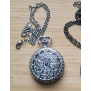 Klocka halsband silverfärgad - Trasig stängning på klockans kedja. Är osäker på om det går att få klockan att fungera, tror att den bara behöver nytt batteri, men har inte öppnat och kollat. Okänt material. Mått i cm, vidd 4, höjd 5,5, kedja 41. 