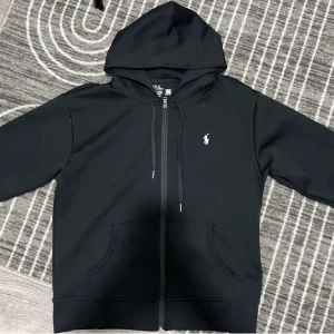 Svart hoodie från Polo Ralph Lauren - Säljer en stilren svart hoodie från Polo Ralph Lauren med dragkedja och det klassiska logotypbroderiet på bröstet. Tröjan har en justerbar huva med snören och praktiska fickor framtill. 