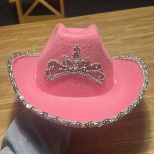 Rosa cowboyhatt - Snygg rosa cowboyhatt med glittrande strassdetaljer och en dekorativ tiara framtill. Hatten har en silverglittrig kant och justerbar hakrem för perfekt passform. Perfekt för att sticka ut på festen!