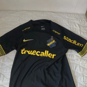 Svart AIK fotbollströja från Nike - Säljer en svart AIK fotbollströja från Nike med gula detaljer och logotyper. Tröjan har korta ärmar och klubbmärket på bröstet. Perfekt för alla AIK-fans! ⚽️