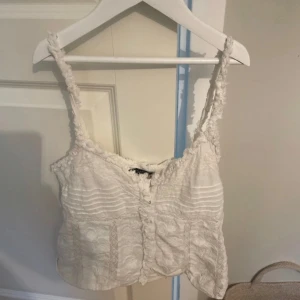 Vit spetslinnetopp - Säljer en söt vit spetslinnetopp med smala axelband och v-ringning. Toppen har fina spetsdetaljer och en lätt ribbad struktur. Perfekt för varma sommardagar eller som ett lager under en kofta.