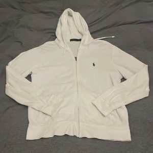 Vit hoodie från Polo Ralph Lauren - Säljer en stilren vit hoodie från Polo Ralph Lauren med dragkedja och det klassiska logotypbroderiet på bröstet. Perfekt för en avslappnad look. Hoodien har långa ärmar och en justerbar huva.