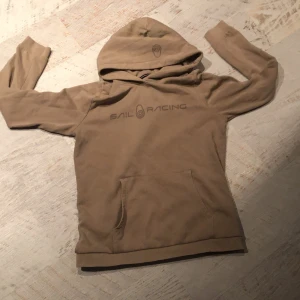 Beige hoodie från Sail Racing - Säljer en beige hoodie från Sail Racing med tryck på bröstet. Den har en klassisk känguruficka och en bekväm huva. Perfekt för en avslappnad stil.