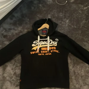 Svart hoodie från Superdry - Säljer en svart hoodie från Superdry med tryck i vitt och orange på framsidan. Den har en justerbar huva med vita snören och en klassisk passform. Perfekt för en avslappnad stil.