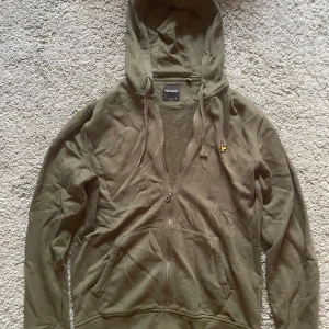 Olivgrön hoodie från Lyle & Scott - Säljer en olivgrön hoodie från Lyle & Scott med dragkedja och huva. Tröjan har en praktisk känguruficka och en liten logotyp på bröstet. Perfekt för en avslappnad stil.