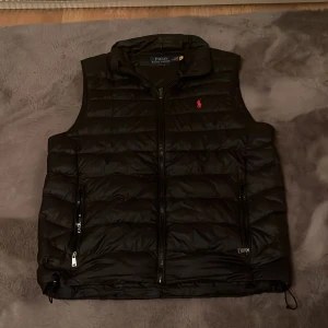 Svart dunväst från Polo Ralph Lauren - Snygg svart dunväst från Polo Ralph Lauren med dragkedja och röd broderad logga på bröstet. Perfekt för lager-på-lager under kyliga dagar. Västen har praktiska fickor med dragkedja och justerbar nederkant. Köpt ifrån care of carl och knappt använd 