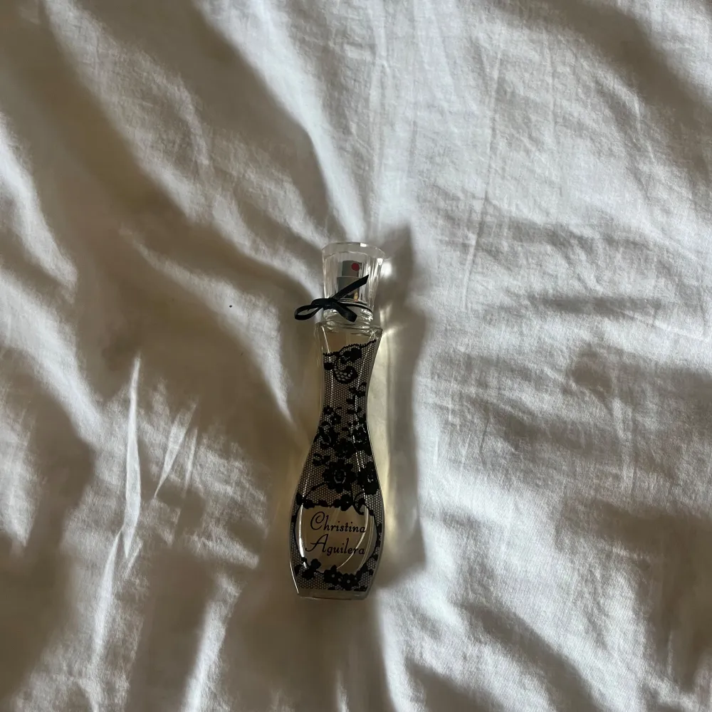 Elegant parfymflaska med svart spetsmönster och en liten svart rosett vid halsen. Flaskan har en smal och kurvig form som ger ett lyxigt intryck. Perfekt för den som vill ha en doft med stil och elegans.. Perfume.
